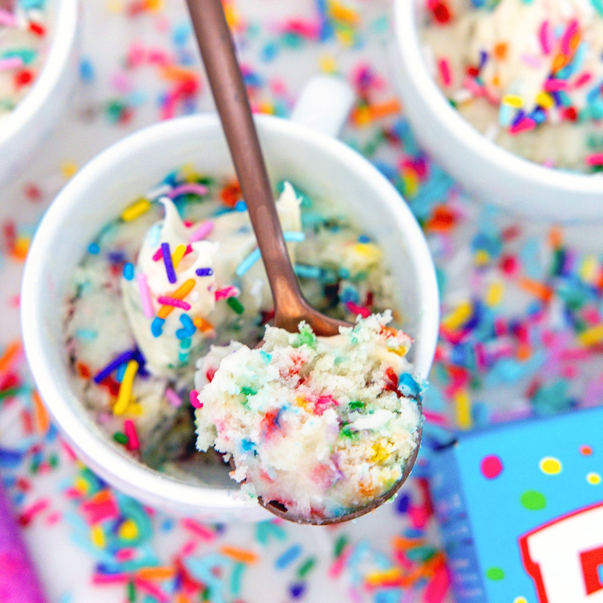 Closeup view of a spoon holding cake over a mug of cake mix mug cake with sprinkles all around.