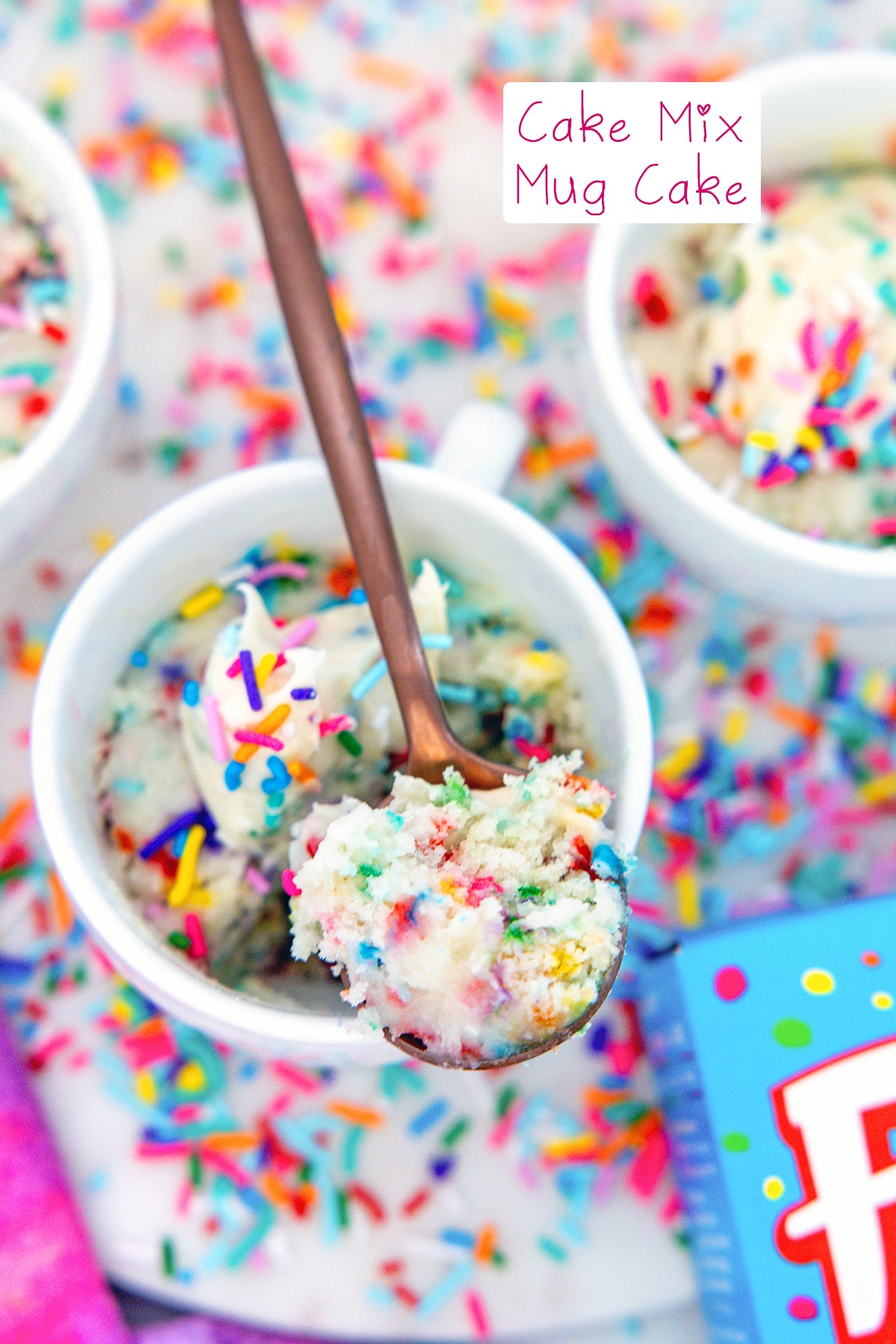 Overhead view of a mug with a spoon and scoop of cake mix mug cake with second mug in background and recipe title at top.