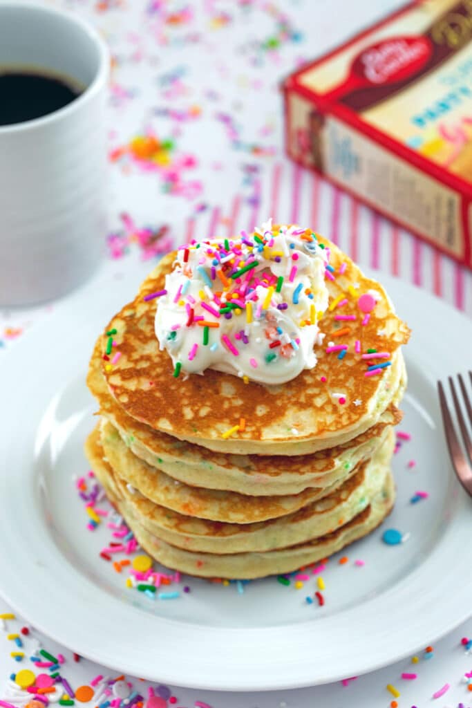 Stack of cake mix pancakes topped with frosting and sprinkles with cake mix box and cup of coffee in background.