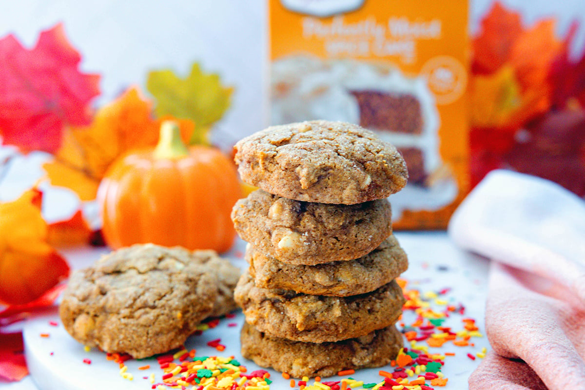 Landscape head-on view of a stack of cake mix pumpkin cookies with sprinkles all around and more cookies and box of cake mix in background.