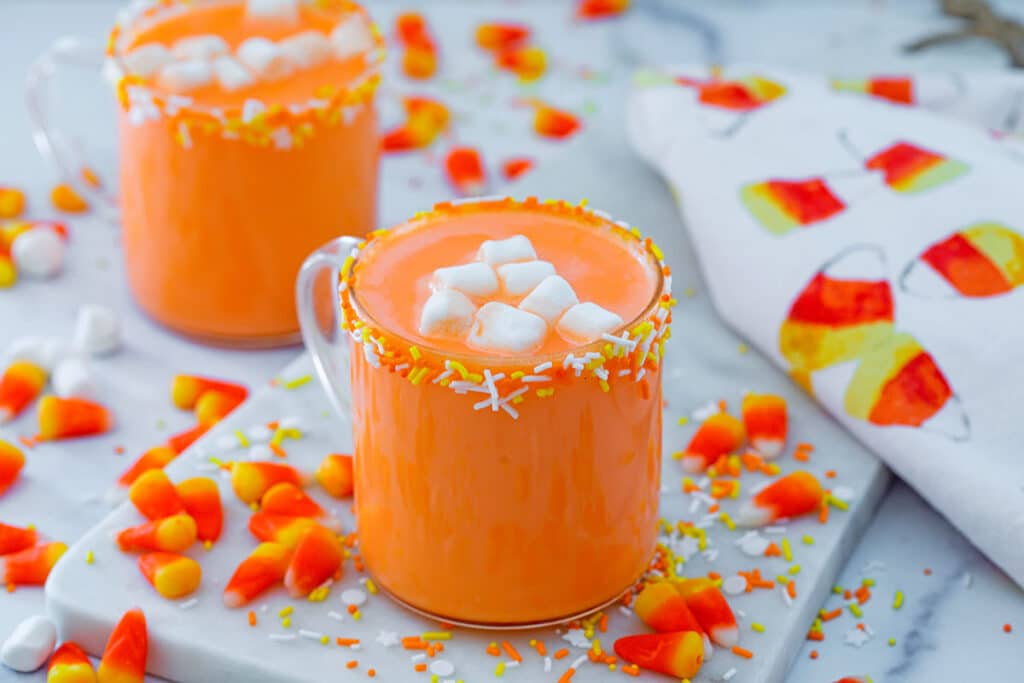 Landscape view of two mugs of candy corn hot chocolate with mini marshmallows on top and candy corn and sprinkles all around.