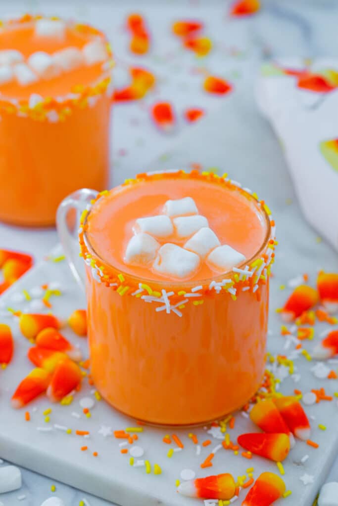 Mug of candy corn hot chocolate with candies and sprinkles all around and second mug in background.