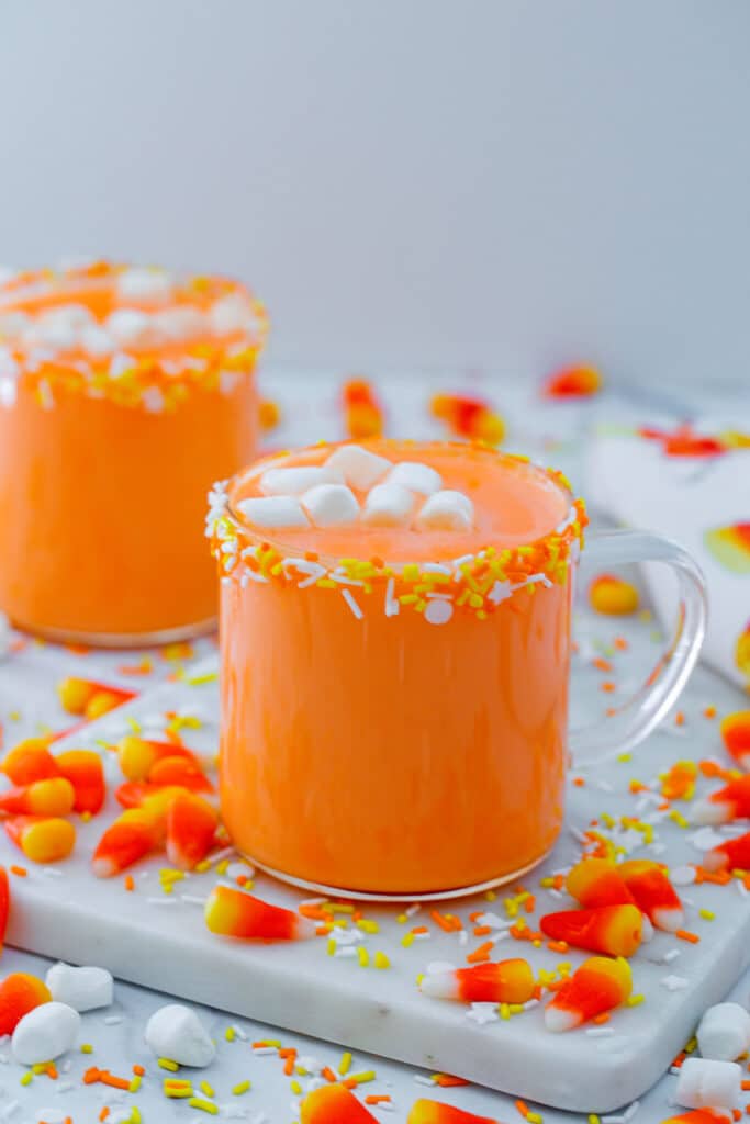 Head-on view of two mugs of candy corn hot chocolate with candy corn and mini marshmallows all around.