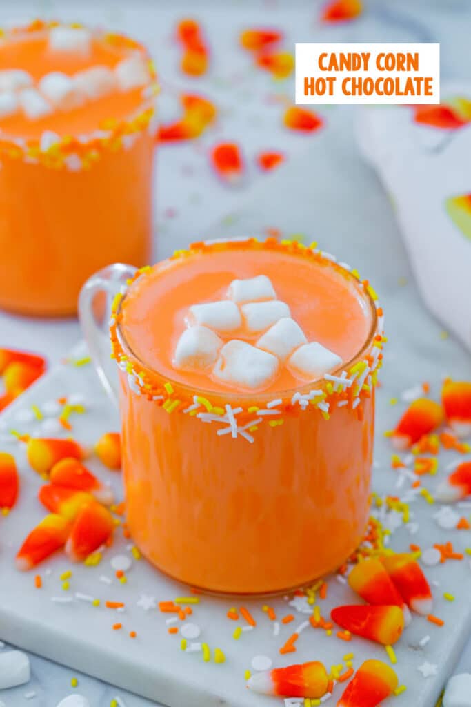 Mug of candy corn hot chocolate with candies and sprinkles all around, second mug in background, and recipe title at top.