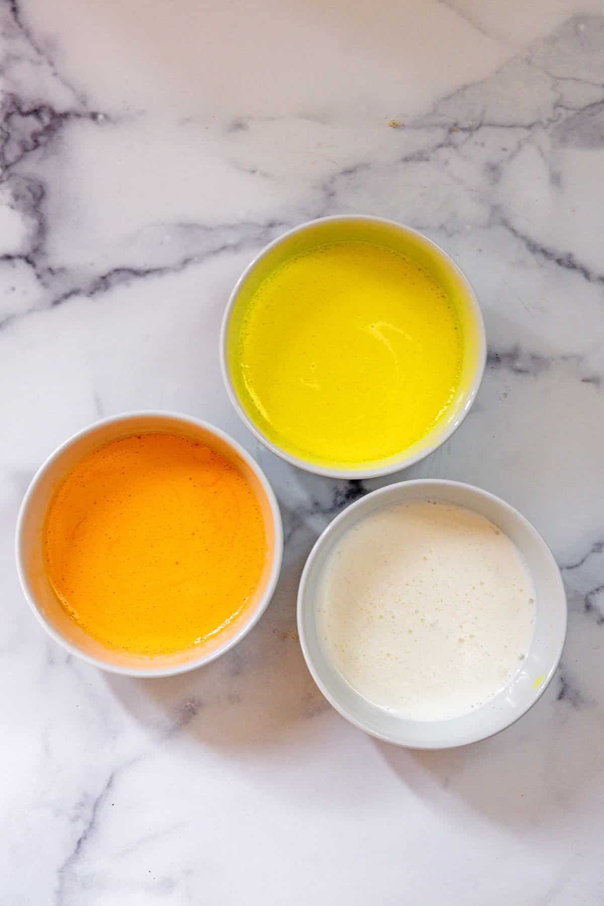 Overhead view of 3 bowls of milkshake mixture colored orange, yellow, and white.