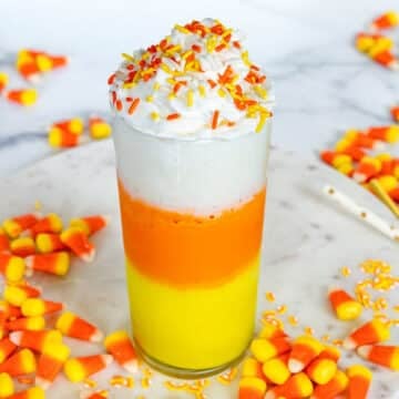 Closeup view of a yellow, orange, and white layered candy corn milkshake topped with whipped cream and sprinkles with candy corns all around.