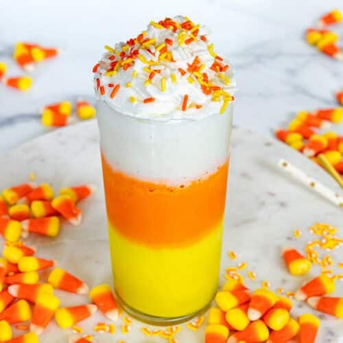 Closeup view of a yellow, orange, and white layered candy corn milkshake topped with whipped cream and sprinkles with candy corns all around.