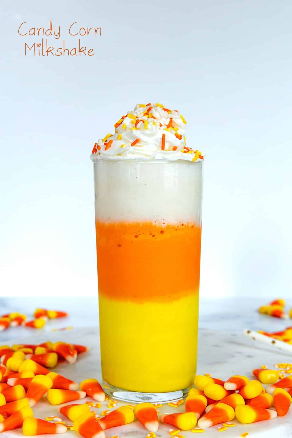 Head-on view of a yellow, orange, and white layered milkshake topped with whipped cream with candy corn all around and recipe title at top.