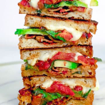Closeup view of caprese grilled cheese sandwich halves with avocado and bacon stacked on top of each other.