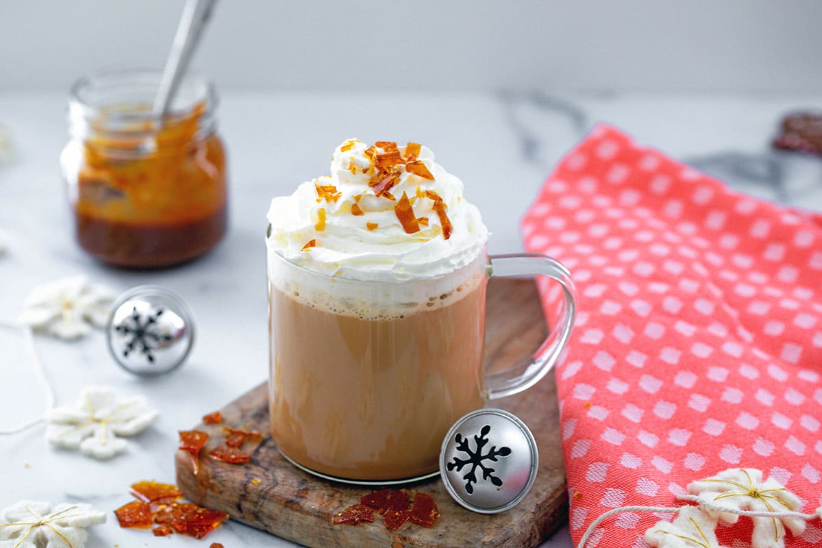 Landscape view of a Caramel Brulée Latte in a clear mug with caramel bits, felt snowflakes, and silver bells all around and jar of caramel sauce in background.
