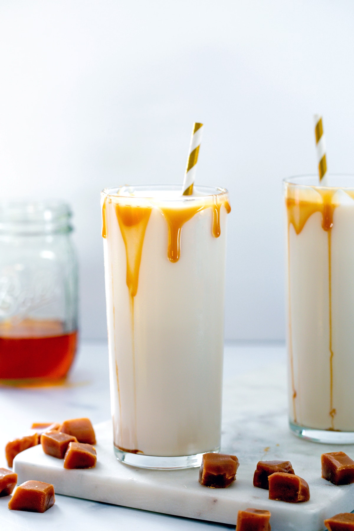 Two glasses of caramel milk with caramel drizzle, caramel syrup in background, and square caramel chews all around.