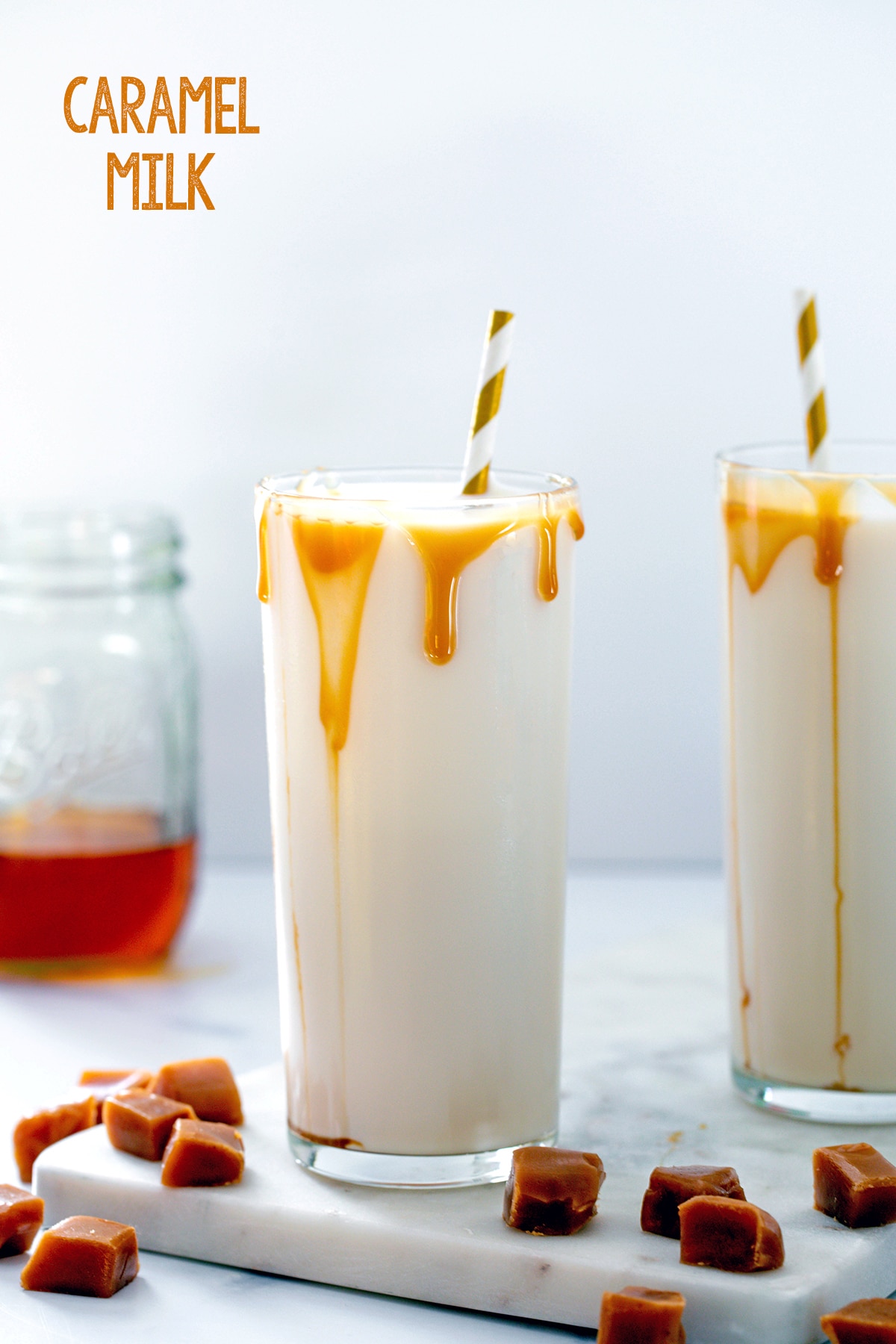 Two glasses of caramel milk with caramel drizzle, caramel syrup in background, and square caramel chews all around with recipe title at top.