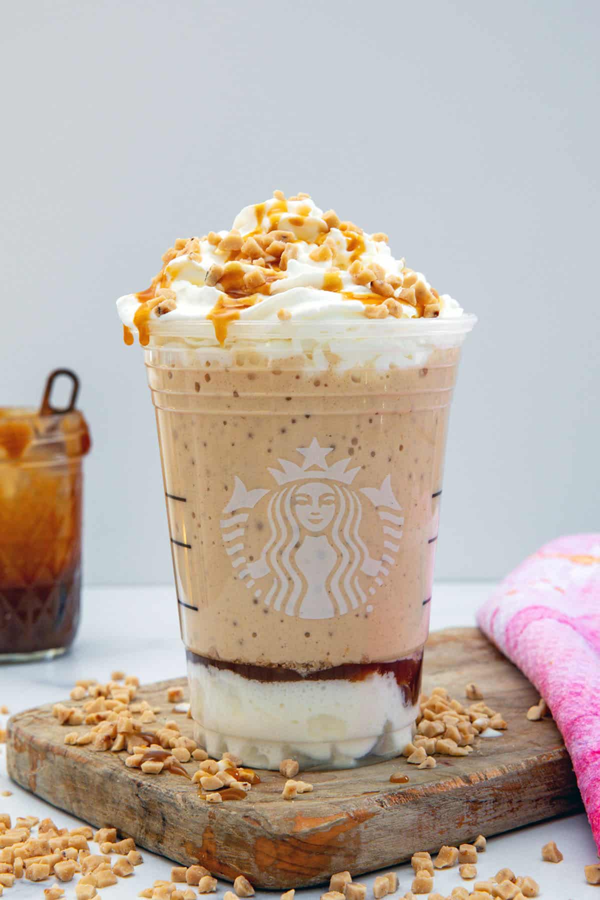 Head-on view of Caramel Ribbon Crunch Frappuccino with caramel pieces all around and jar of caramel sauce in background.