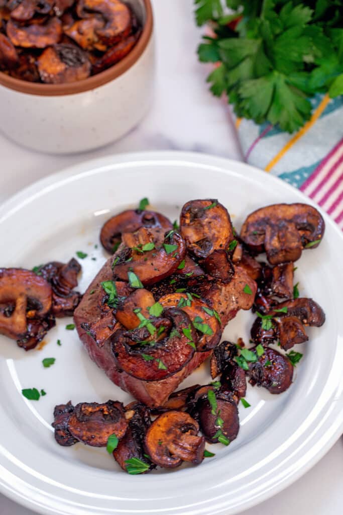 Caramelized mushrooms on top of steak on white plate with bunch of parsley in the background.