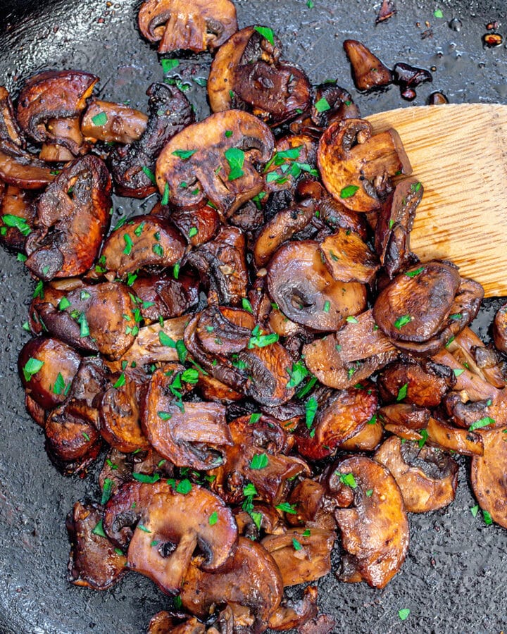 Closeup view of caramelized mushrooms in cast iron skillet with wooden spatula.
