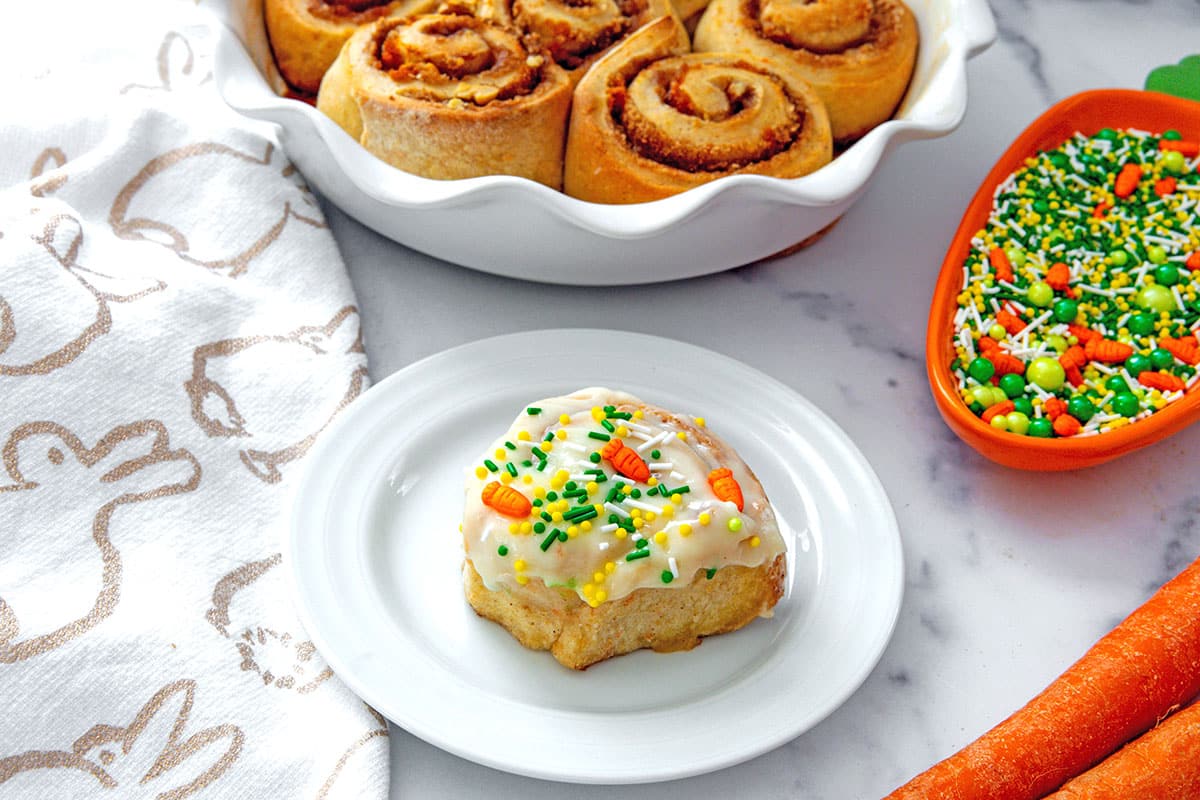 Landscape view of carrot cake cinnamon rolls with carrot-shaped bowl of sprinkles and fresh carrots.