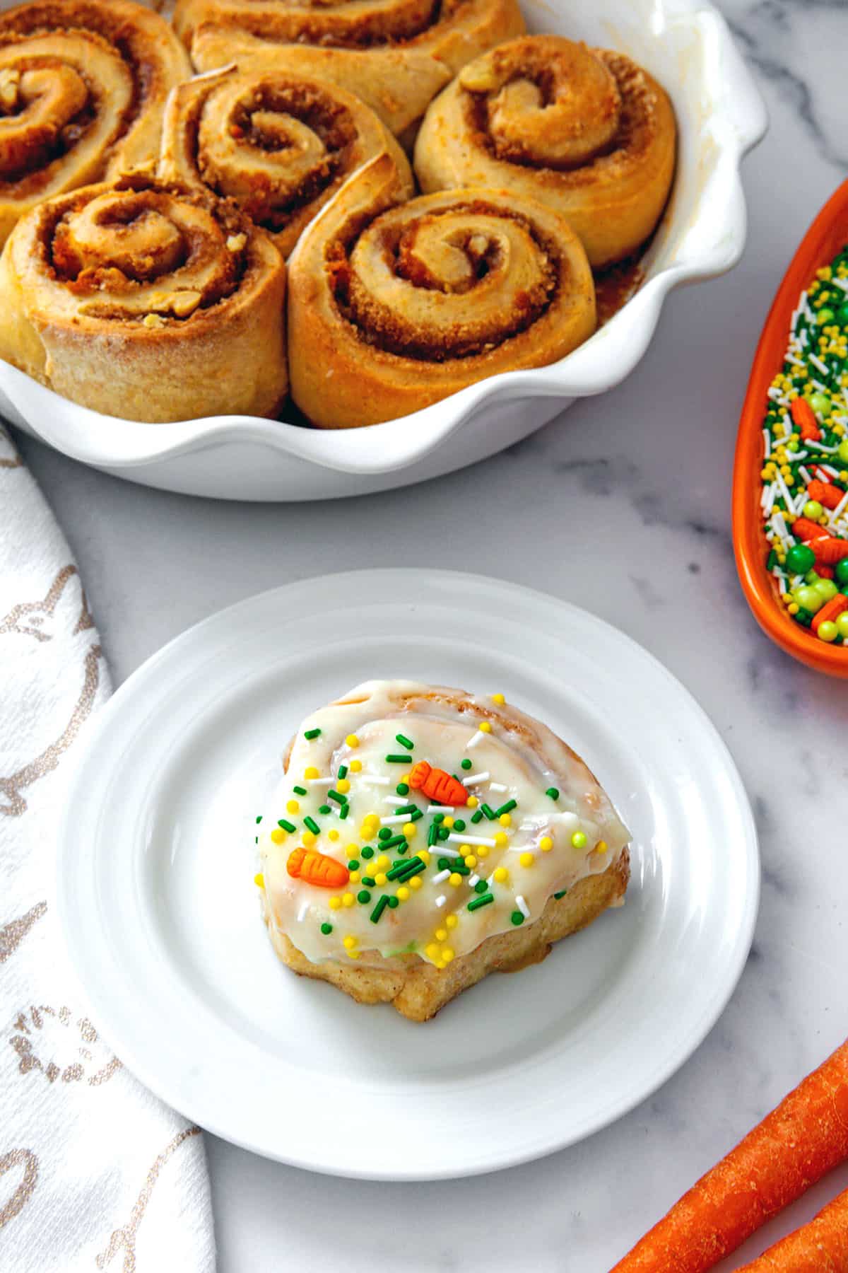 Cream cheese frosted carrot cake cinnamon roll on a plate with dish of bare cinnamon rolls in background.