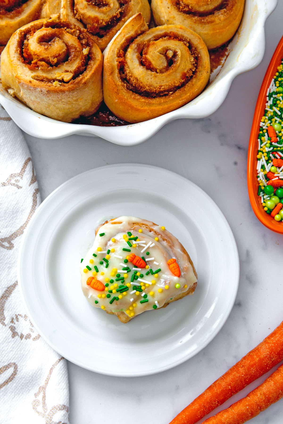 Bird's eye view of a carrot cake cinnamon roll on a white plate with platter of rolls in background with whole carrots and bowl of sprinkles.