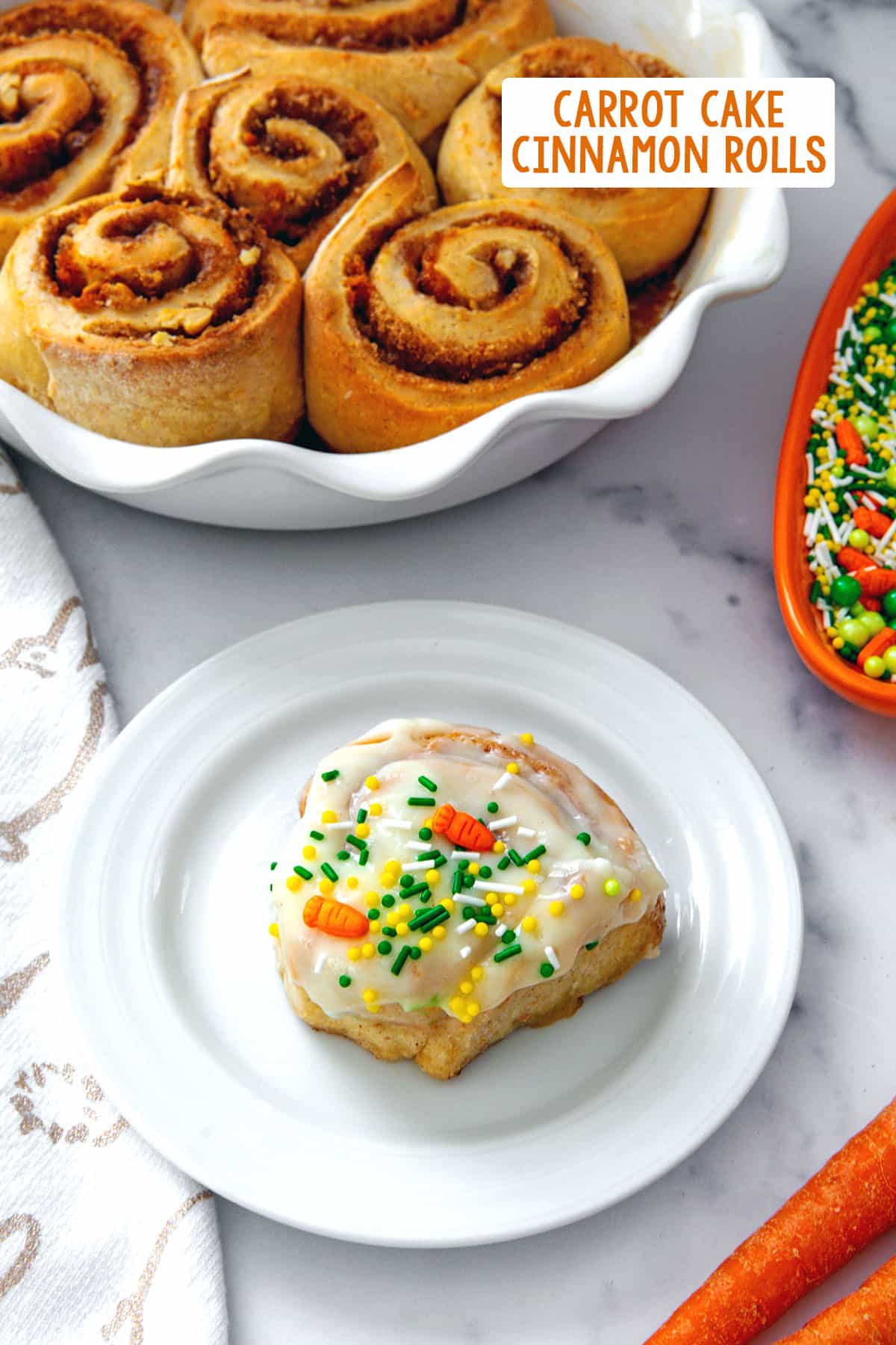 Cream cheese frosted carrot cake cinnamon roll on a plate with dish of bare cinnamon rolls in background and recipe title at top.