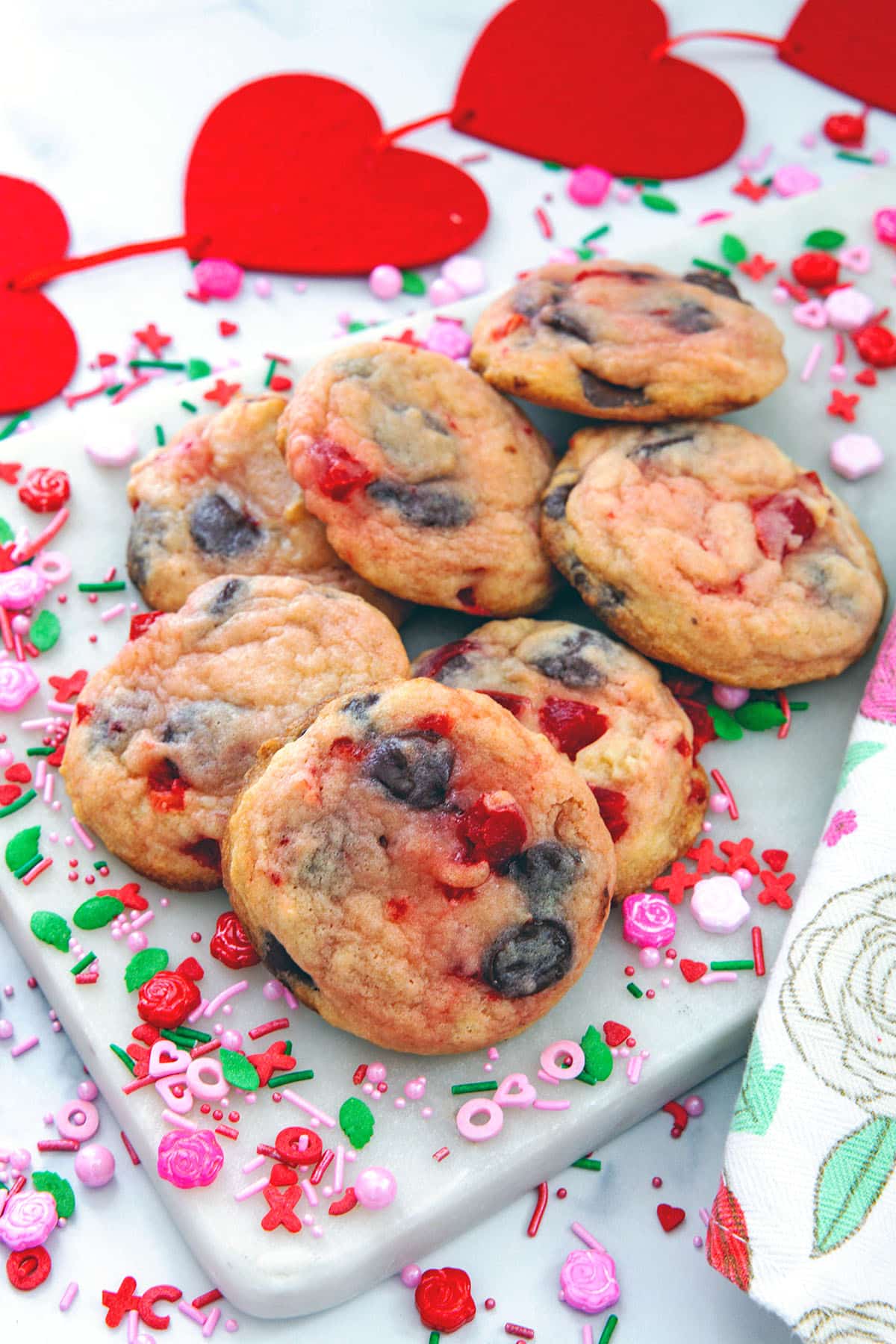 Cherry chocolate chip cookies on a marble surface with Valentine's Day sprinkles all around and red hearts in backgorund.