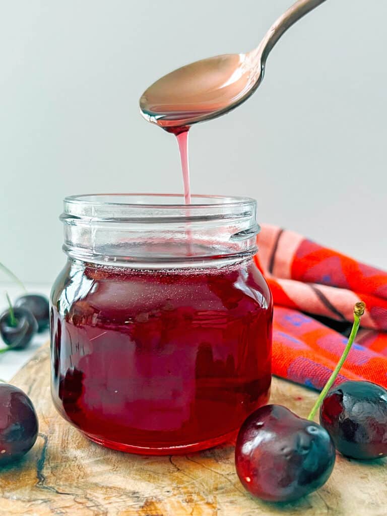 Small jar of cherry syrup with spoon drizzling syrup off of it.