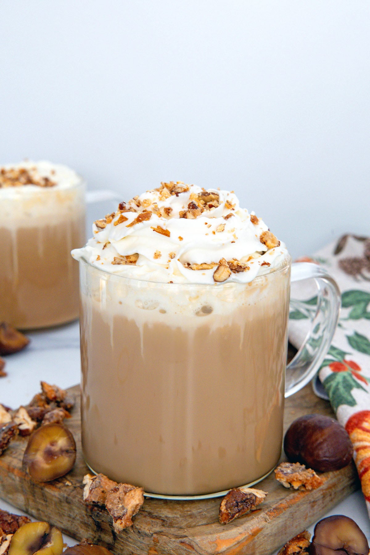 Head-on view of a Chestnut Praline Latte with chestnuts and praline all around, second drink in background.