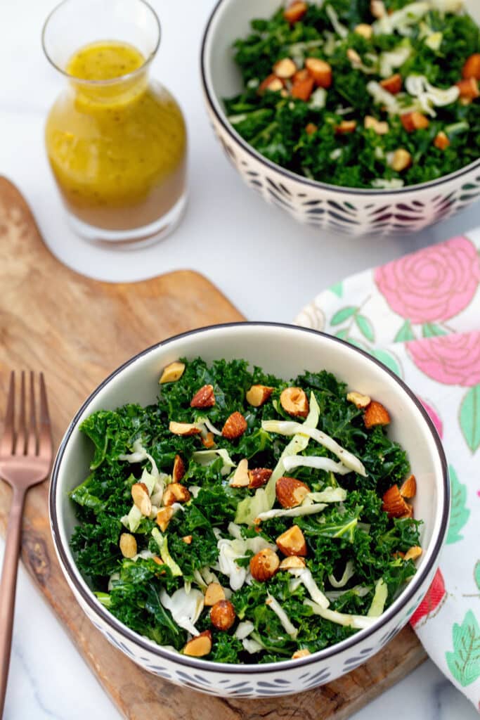 Kale salad in a bowl with almonds with second bowl of salad and small carafe of dressing in background.