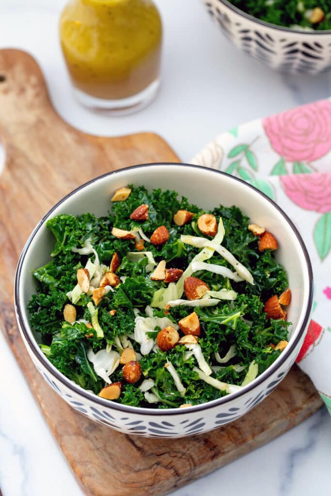 Overhead view of a kale salad with green cabbage and almonds with small bottle of dressing in background.