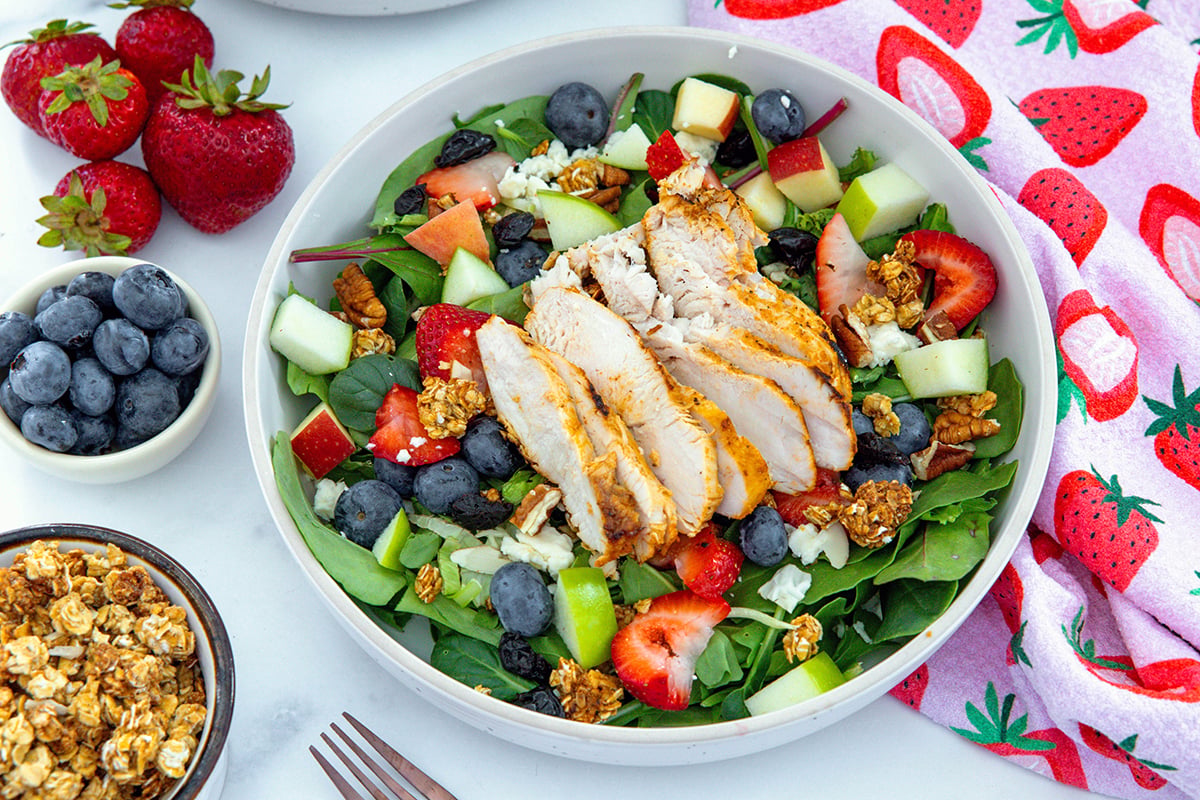 Landscape overhead view of Chick-Fil-A market Salad with grilled chicken on top and bowl of granola and blueberries on the side with strawberries.