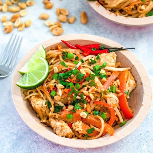 Closeup view of a bowl of chicken pad Thai with Thai chili peppers and lime wedges and peanuts in the background.