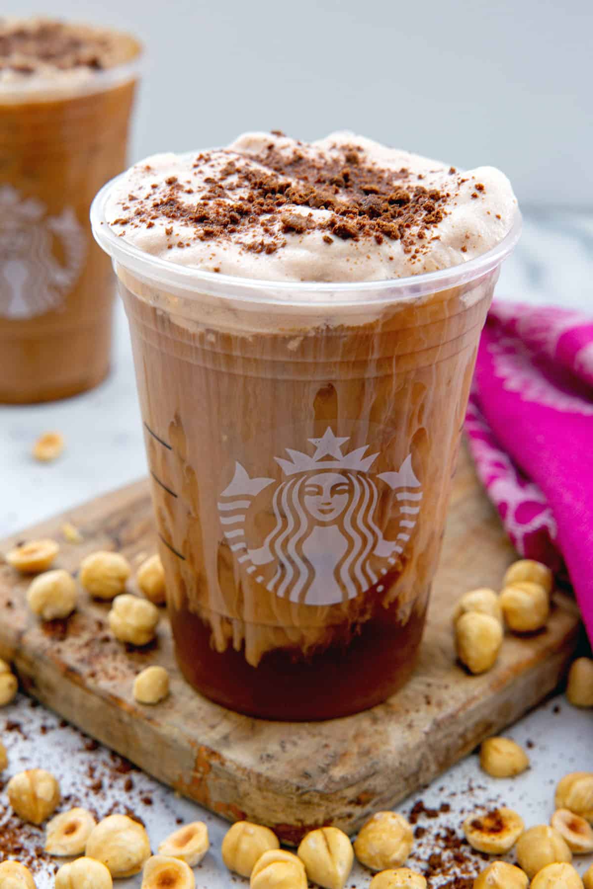 Chocolate Hazelnut Cookie Cold Brew in a Starbucks cup with cold foam and cookie crumbles on top and hazelnuts all around.