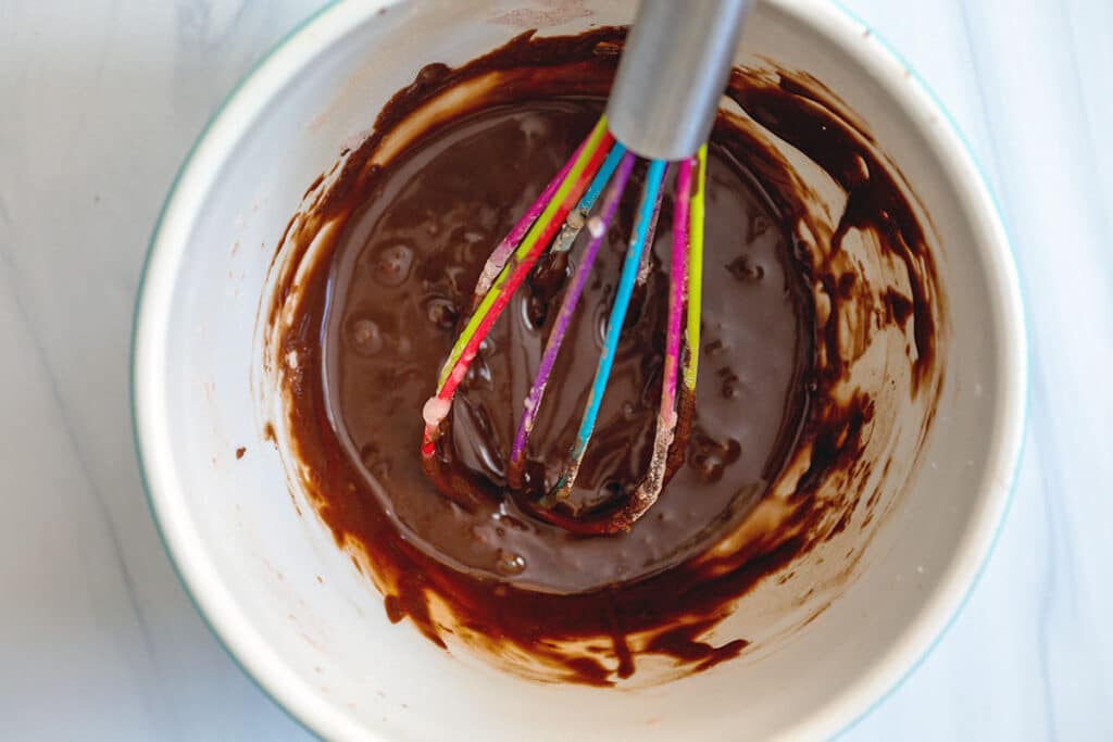 Chocolate icing in bowl with rainbow whisk.