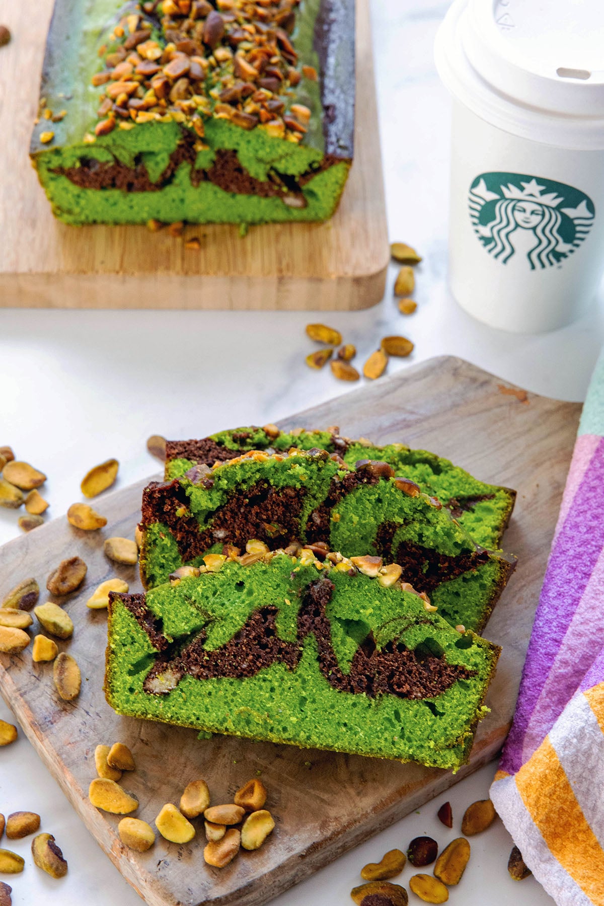 Two slices of chocolate pistachio loaf on a wooden board with pistachios all around and loaf in background.