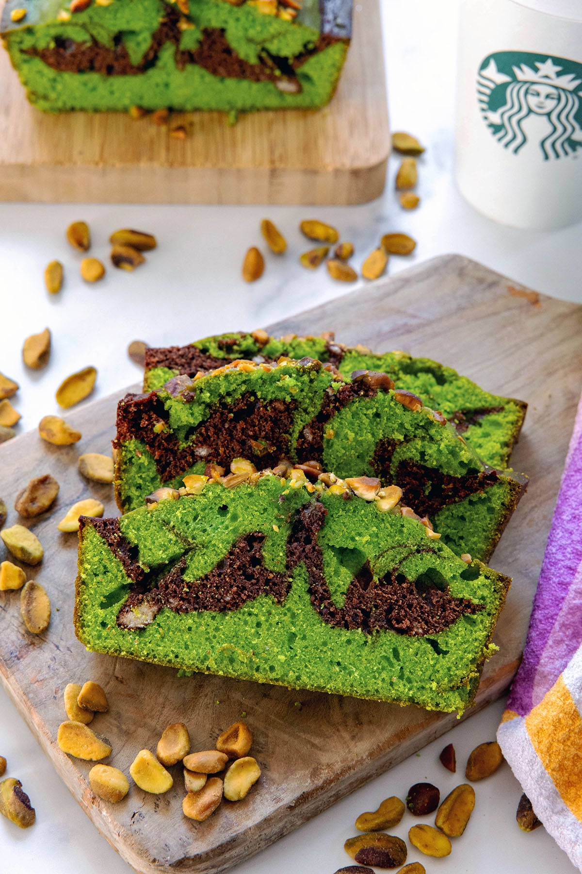 Overhead view of slices of chocolate pistachio loaf on a wooden board with loaf and Starbucks coffee cup in background.
