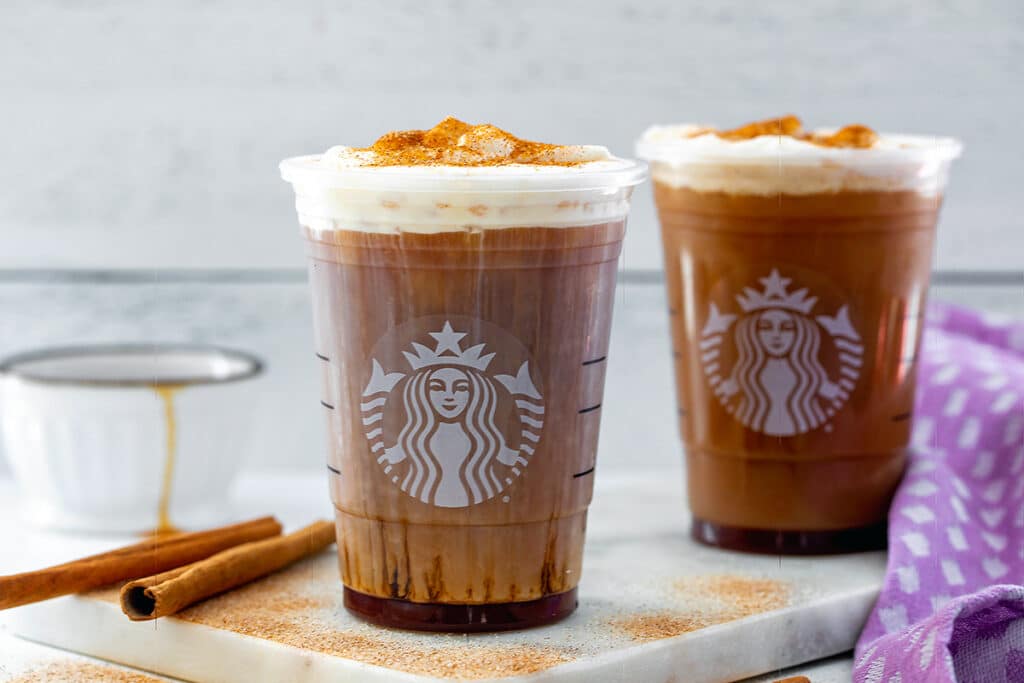Landscape head-on view of two Starbucks cups with cinnamon caramel cream cold brew with cinnamon sticks and cup of caramel in the background.