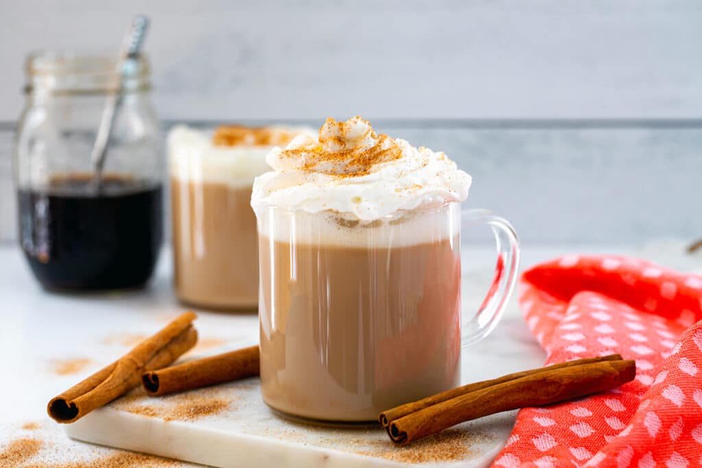 Head-on landscape view of a cinnamon dolce latte with whipped cream with second latte and jar of syrup in background and cinnamon sticks all around.