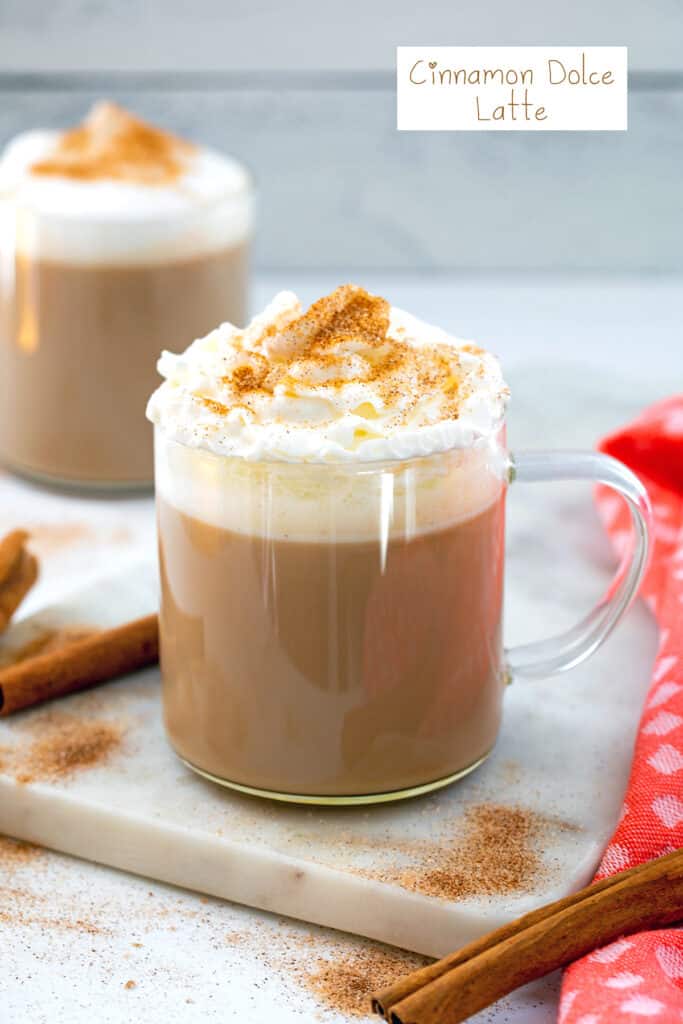 Head-on view of a cinnamon dolce latte with whipped cream and ground cinnamon with cinnamon sticks on the side and recipe title at top.