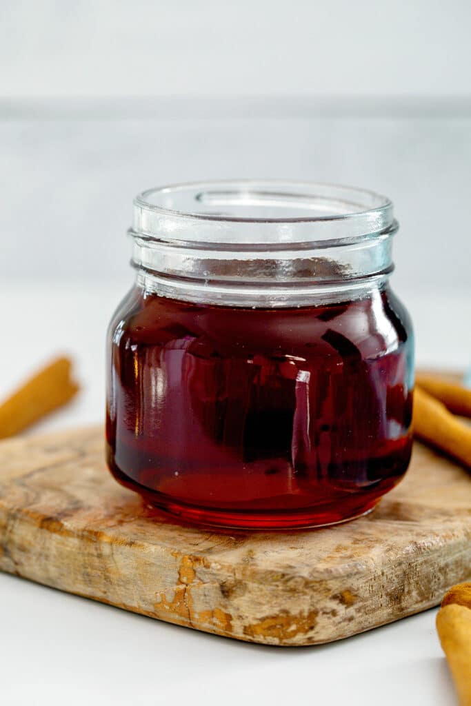 Small mason jar of cinnamon dolce syrup on a wooden board with cinnamon sticks all around.