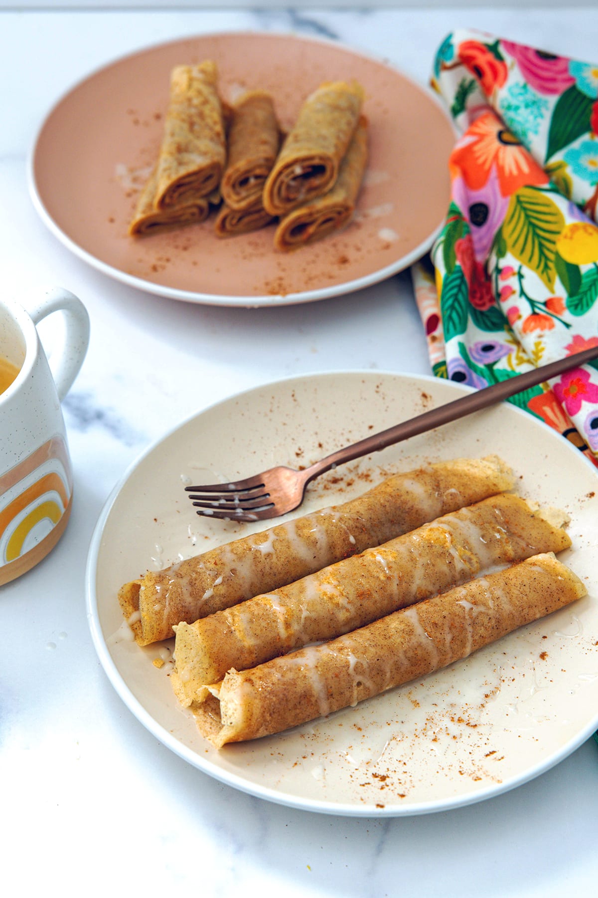 Two plates of rolled up cinnamon roll crepes with mug of coffee.