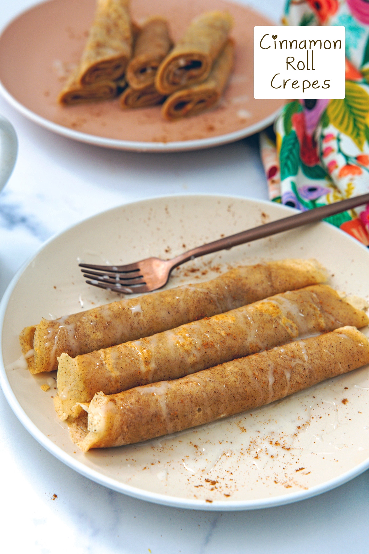 Plate of rolled up cinnamon roll crepes topped with icing with another plate in background and recipe title on top.