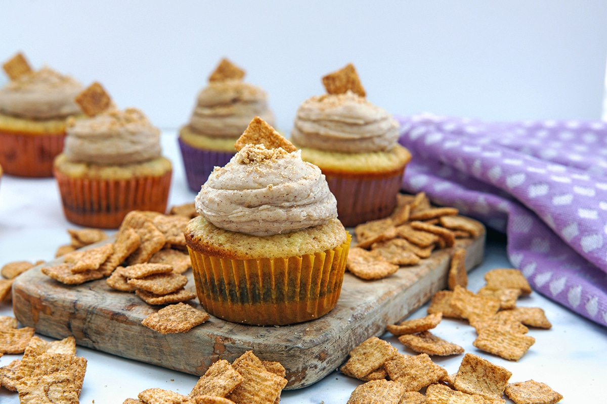 Landscape head on view of multiple Cinnamon Toast Crunch cupcakes on a wooden board with cereal all around.