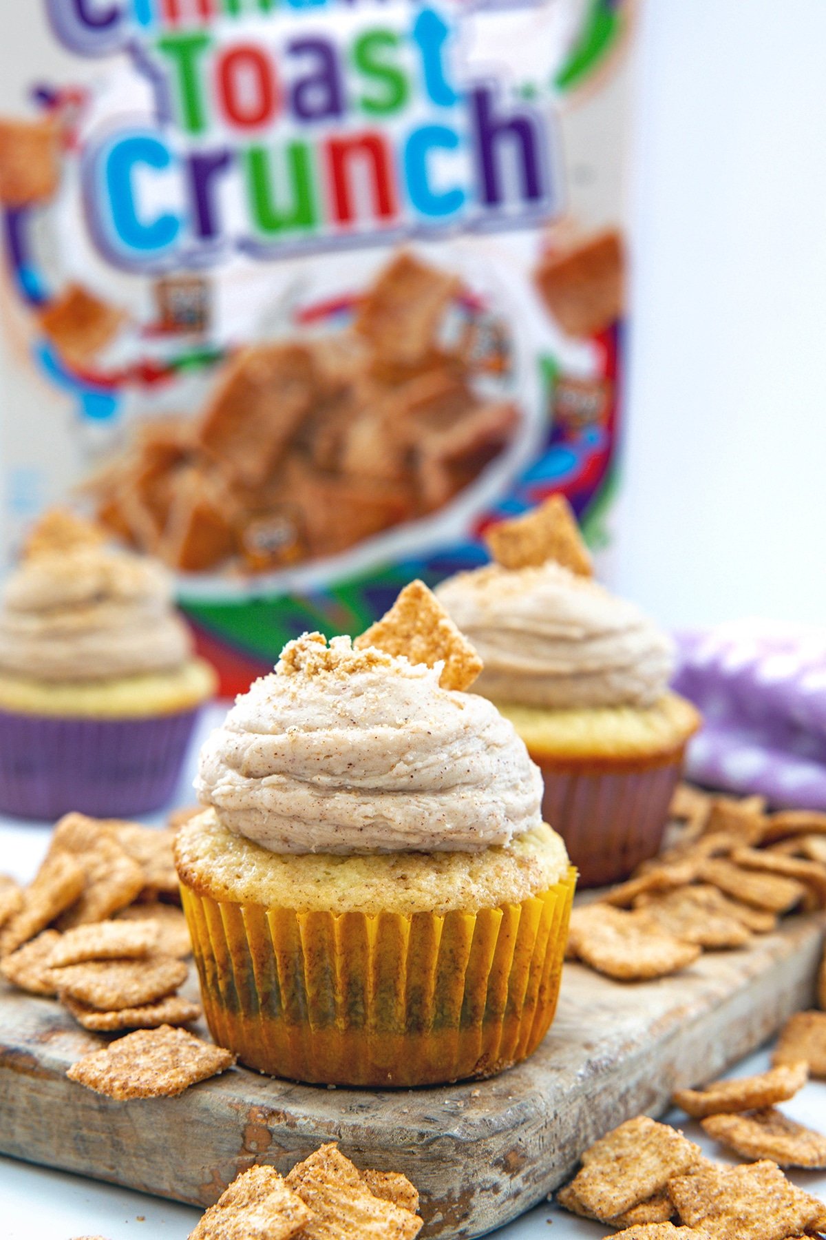 Head-on view of a Cinnamon Toast Crunch cupcake with cereal all around and a cereal box in the background.