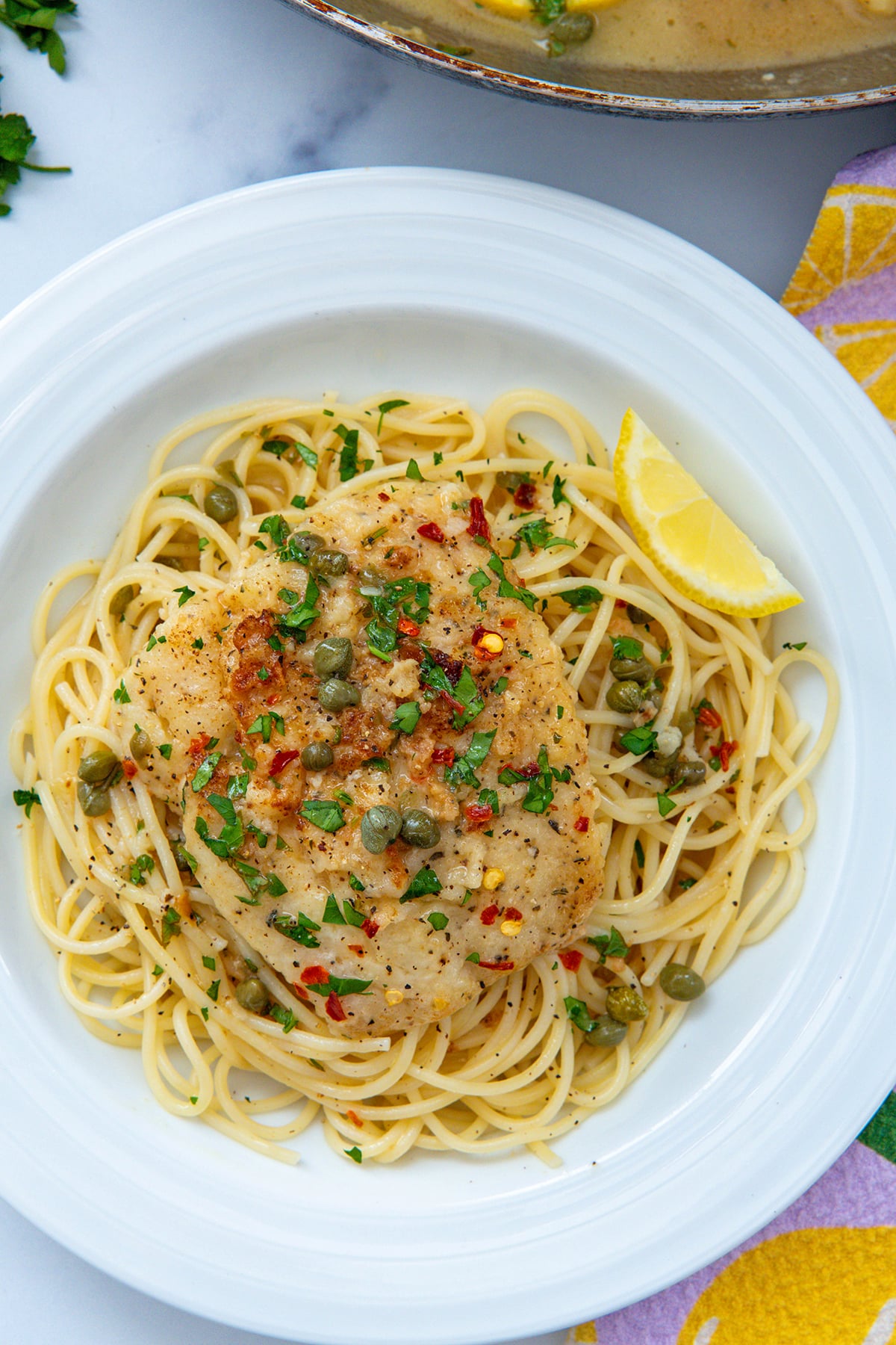 Overhead closeup view of a plate with linguine topped with cod piccata with capers and lemon wedge.