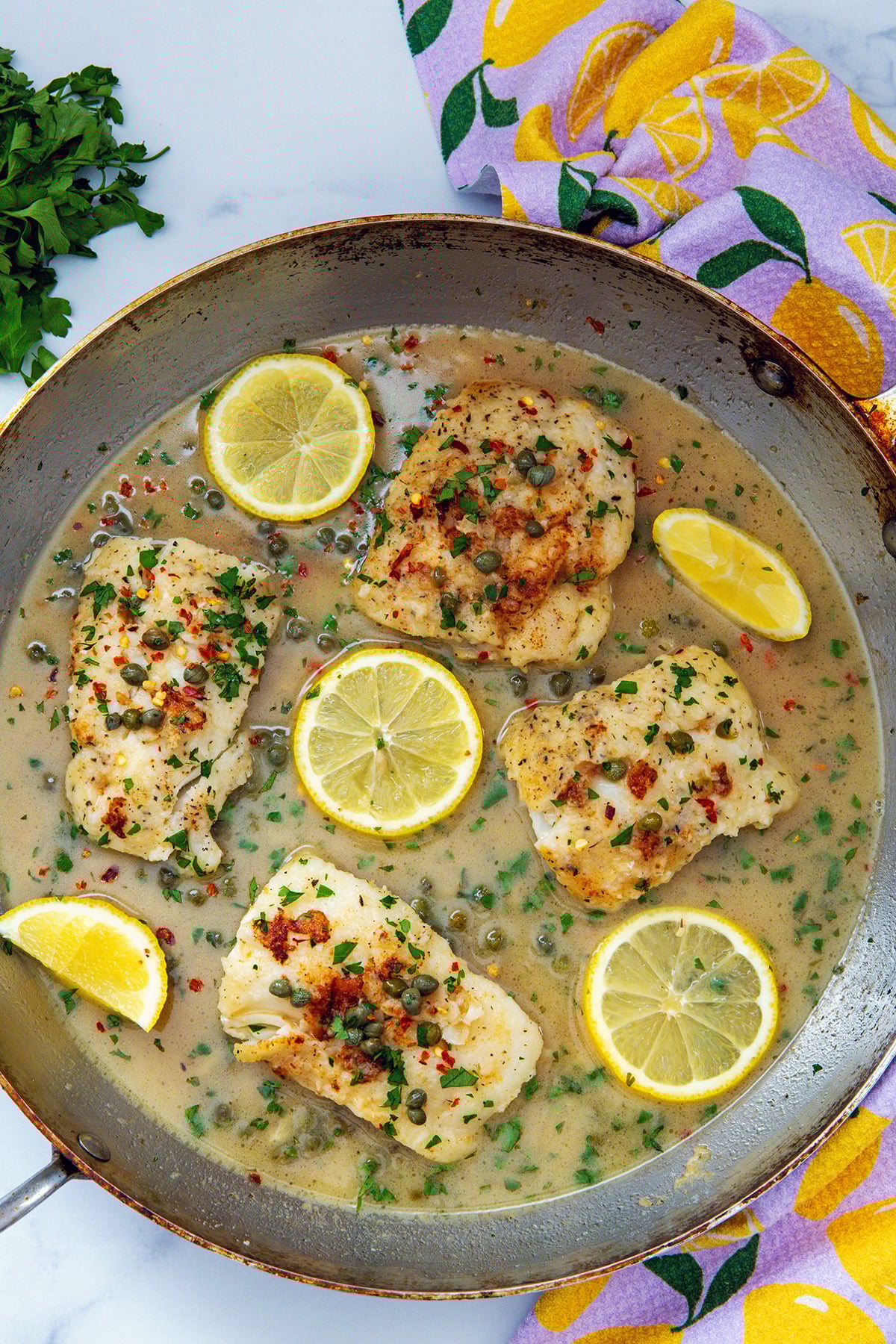 Overhead view of a skillet with golden cod filets, piccata sauce, and lemon slices.