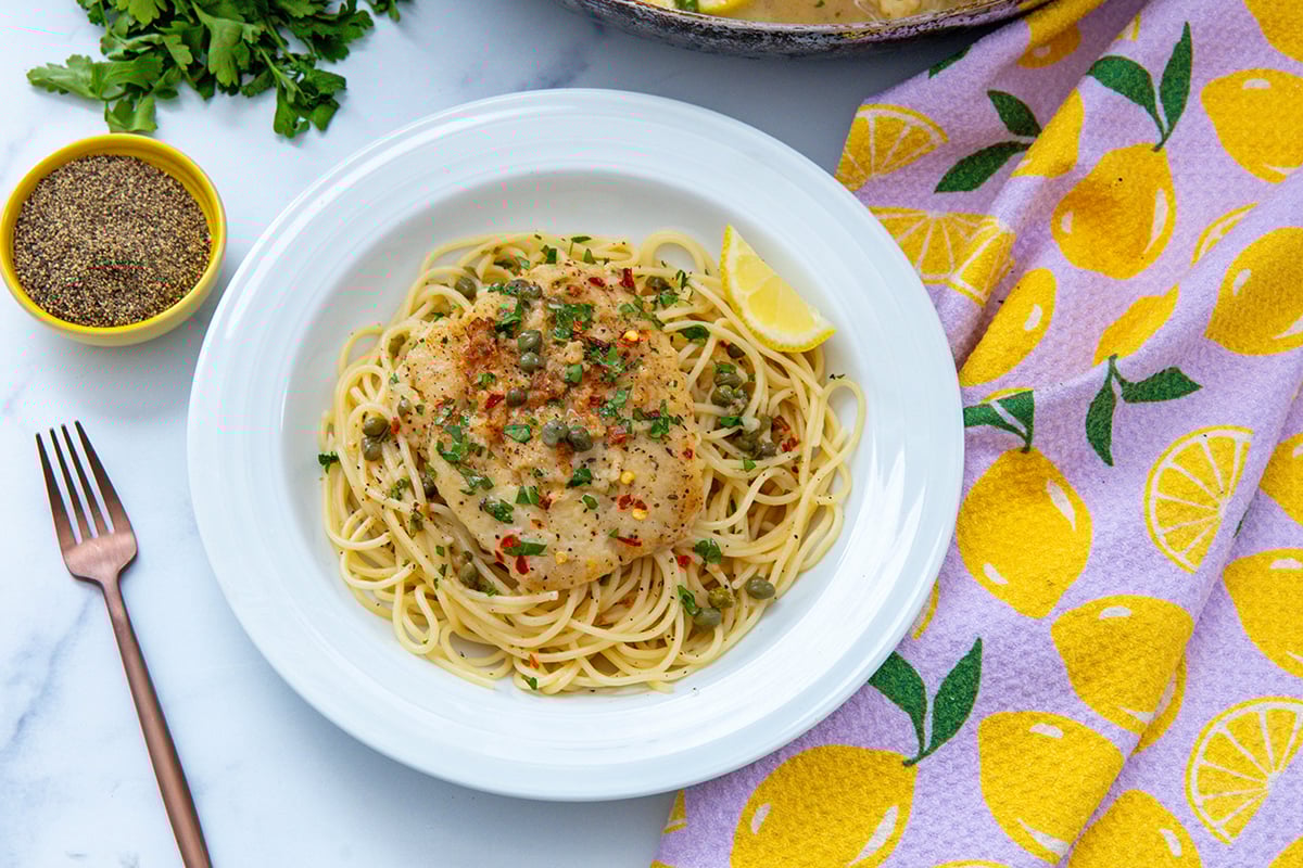 Landscape overhead view of cod piccata over linguine with parsley and black pepper in background.