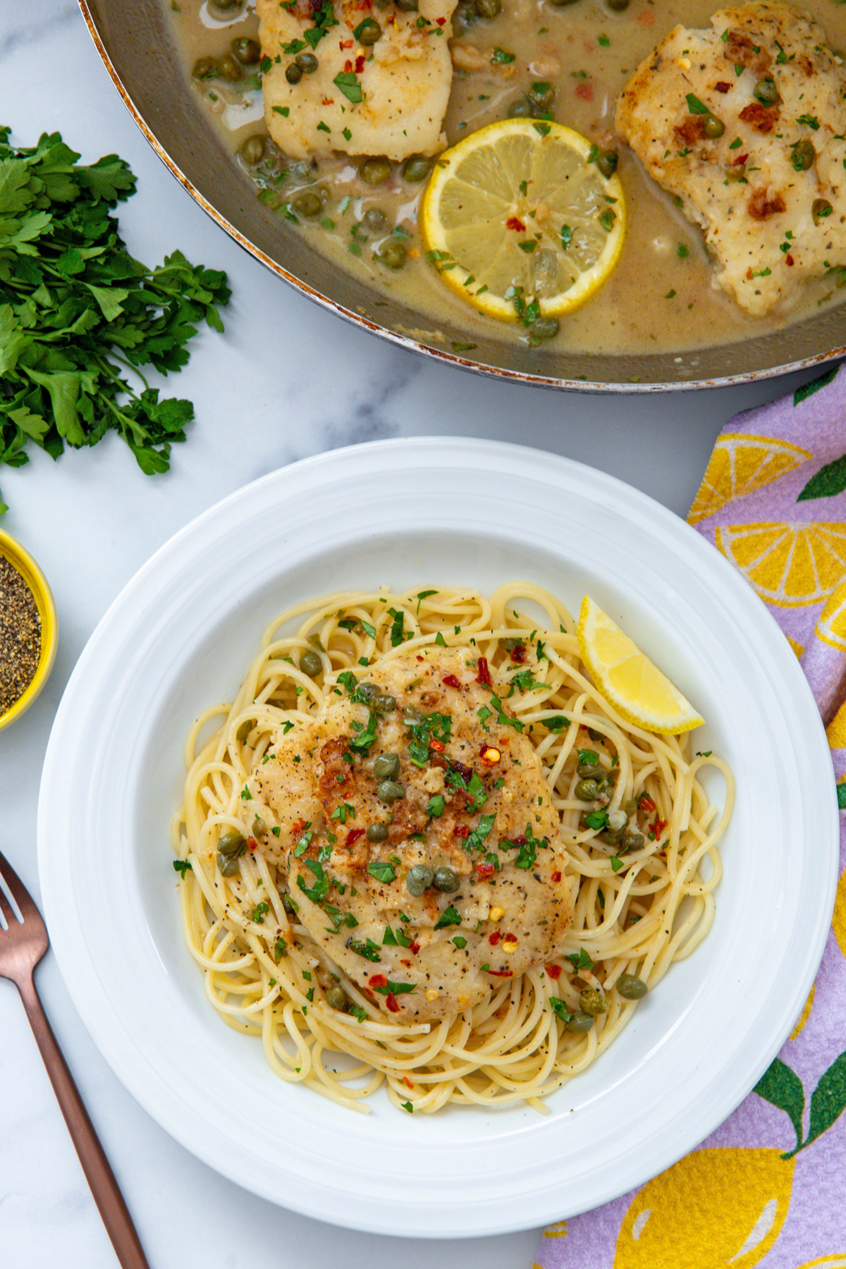 Overhead view of cod piccata served over pasta with skillet of lemon cod in background.