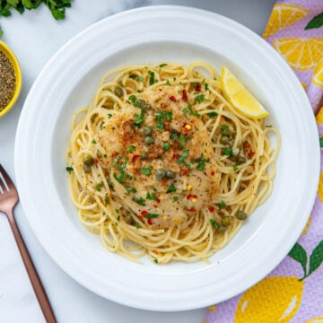 Closeup view of cod piccata over pasta with capers and lemon wedge.