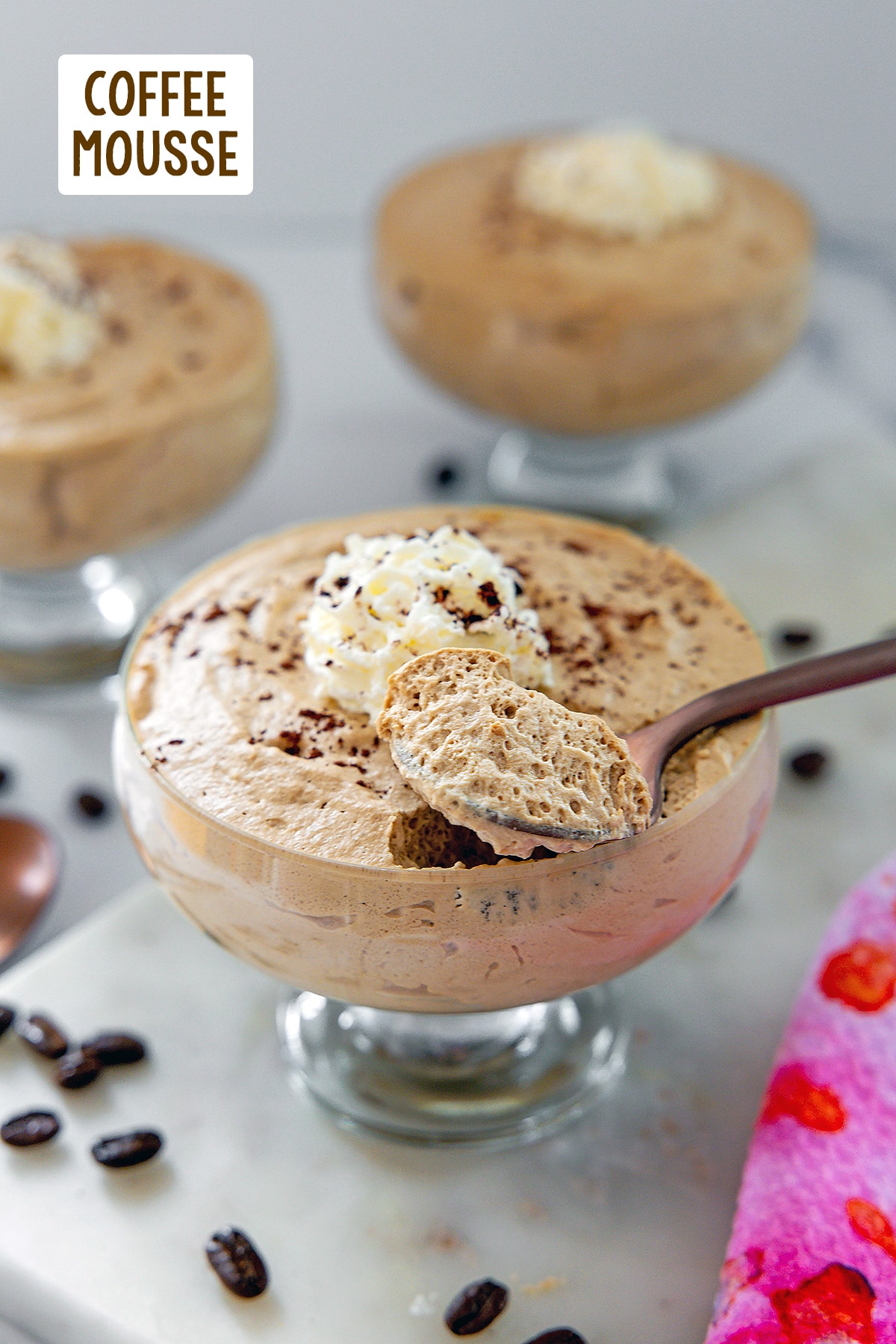 Glass ramekin of coffee mousse with whipped cream and spoon with more ramekins and coffee beans in background with recipe title at top.