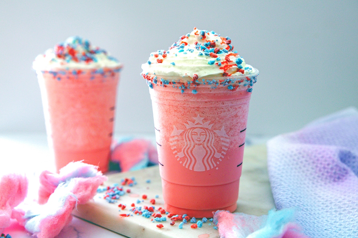 Head-on landscape view of two Starbucks cups with Cotton Candy Frappuccinos with whipped cream and pink and blue sprinkles.