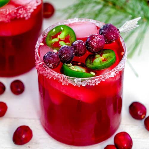 Closeup view of a cranberry jalapeño margarita with cranberries all around and holiday holly in background.